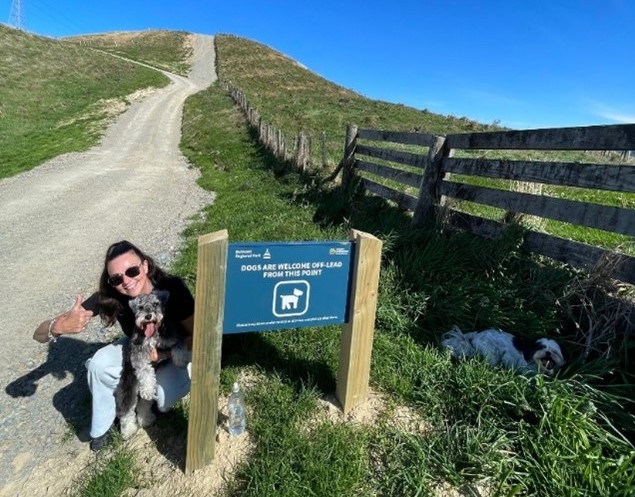 A woman crouches with her dog next to a "dogs are welcome off-lead beyond this point" sign